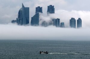 짙은 해무가 해안 도시를 덮으며 바다에서 육지로 천천히 밀려오는 모습. 건물과 다리가 흐릿하게 잠겨 있고, 하늘과 바다의 경계가 모호해진 장면.